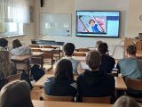 Klasa szkolna. Kilkunastu uczniów siedzi przodem do tablicy multimedialnej, biorą udział w lekcji online dotyczącej...