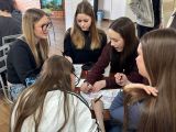 Pięć uczennic siedzi przy stoliku na szkolnej auli, kolorują obrazki, są uśmiechnięte.