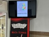 Telewizor na ścianie, na nim wyświetlany obraz kolorowego mózku z napisem Europejski Dzień Mózgu 18 marca.Pod telewizorem...