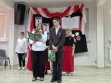 Grupa uczniów w strojach galowych stoi w zadumie. W pierwszym rzędzie dziewczyna z mikrofonem w ręku oraz chłopak w czarnym...