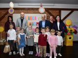 Na pierwszym planie grupa uśmiechniętych dzieci. Na drugim planie grupa dorosłych osób w odświętnych strojach