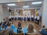 Uczniowie siedząc na krzesłach obserwują występ uczniów ubranych w stroje ludowe