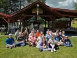 Grupa uśmiechniętych uczniów siedzi na soczystej, zielonej trawie. Za nimi duży piec do wypiekania chleba okryty dachem