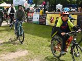 Grupa osób na rowerach wjeżdża na plaży pod scenę. Wśród rowerzystów są rodośli i dzieci. Niektóre osoby mają kaski na...