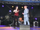 Na scenie mężczyzna w jasnym garniturze wręcza dziewczynie szare pudełko z nagrodą.