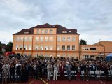 Przed budynkiem szkoły na krzesłach siedzi tłum ludzi (około 400 osób).