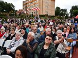 Na krzesłach w kilkunastu rzędach siedzą osoby w różnym wieku. W tle pomarańczowy budynek.