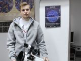 W pomieszczeniu stoi uczeń liceum w szarej bluzie z kapturem, z blond włosami. Przed nim stoi teleskop.