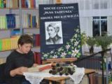 Kobieta z krótkimi włosami siedzi przy stoliku i czyta książkę. W tle sztaluga ze zdjęciem mężczyzny.