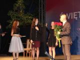 Na scenie stoją trzy uczennice liceum obok mężczyzny w garniturze, który trzyma duży bukiet czerwonych róż. W dali drugi...
