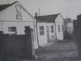 Czarno-białe zdjęcie, parterowy budynek ze spadzistym dachem. Nad wejściem widoczny szyld z nazwą szkoły.