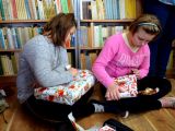 Dwie uczennice szkoły podstawowej siedzi na podłodze i rozpokowuje prezenty. W tle regały z książkami.