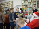 Grupa uczniów szkoły podstawowej stoi przed św. Mikołajem i odbiera prezenty. W tle regały z książkami.