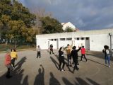 Siedmioro uczniów liceum i dwie kobiety grają w piłkę ba boisku asfaltowym. W tle biały, niski budynek.