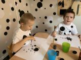 Dwóch chłopców z klasy I-III w białych koszulkach w czarne kropki siedzi przy stoliku i maluje kropki.
