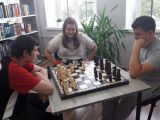 Troje uczniów klasy VII siedzi przy stoliku i gra w szachy. W tle duży, zielony kwiat w donicy.