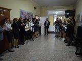 Nauczyciele stoją w rzędzie przy ścianie na korytarzu. Przed nimi stoi grupa uczennic liceum i śpiewa do mikrofonów.