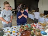 Uczniowie szkoły podstawowej wraz z nauczycielami oglądają wystawę. Na stołach leżą kolorowe wyroby z gliny.