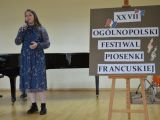 Dziewczyna w niebieskiej sukience z mikrofonem, za nią stoi czarny fortepian, po prawej stronie tablica na sztaludze.