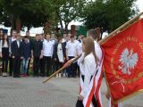 Dwójka uczniów w białych koszulach ze sztandarem na tle grupy uczniów liceum, u góry zdjęcia widoczne korony drzew.