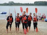 Pięciu licealistów w kamizelkach ratunkowych stoi na plaży, trzymajc wiosła. W tle jezioro.