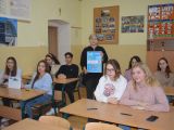 Uczniowie liceum siedzą w ławkach szkolnych w sali lekcyjnej. Między ławkami chodzi kobieta, która trzyma przed sobą plakat.
