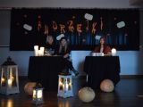 Trzy licealistki siedzą za dwoma stolikami. Na stolikach stoją zapalone świece. Przed stolikami leżą dynie i stoją lampiony.