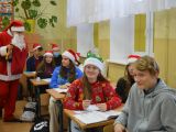 Uczeń w stroju św. Mikołaja chodzi po klasie i częstuje licealistów cukierkami. Uczniowie siedzą w ławkach szkolnych.