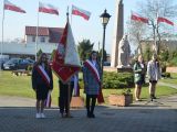 Troje uczniów liceum stoi na dziedzińcu szkoły. Uczeń w środku trzyma przed sobą sztandar.