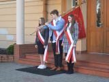 Treoje uczniów liceum wychodzi ze szkoły. Chłopak w środku niesie na ramionach sztandar.