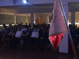 Uczniowie liceum i nauczyciele siedzą na widowni. Po prawej stronie stoi opaty na stojaku sztandar szkoły.