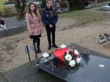 Dwoje licealistów stoi przed pomnikiem na cmentarzu. Na płycie pomnika kwiaty i znicz.