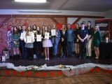 Dziewiętnaście osób soti na scenie w dwóch rzędach.  Niektóre osoby trzymają w rękach dyplomy.