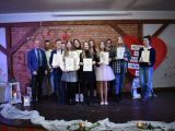 Na scenie stoi kilka osób, trzymają dyplomy i nagrody. Po lewej stronie stoi mężczyzna w niebieskiej  marynarce.