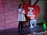 Uczennica szkoły podstawowej w białej sukience stoi na na scenie, w ręku trzyma mikrofon.