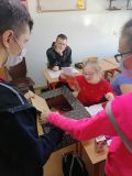 Uczennica szkoły podstawowej w okularach siedzi w ławce, wyciąga z pudełka kartkę. Wokół niej stoi troje innych uczniów.
