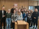 Uczniowie liceum stoją w grupie w sali sądowej. Z lewej strony stoi mężczyzna, z prawej kobieta. Przed nimi stoi mównica.