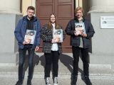 Troje uczniów liceum stoi obok siebie na schodach, przed wejściem do szkoły. Wszyscy trzymają  przed sobą dyplom  książką.