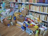 Na podłodze w bibliotece leży duża sterta produktów spożywczych i środków higienicznych. W tle regały z książkami.