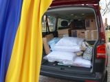 Otwarty bagażnik busa. W bagażniku kartony i białe poduszki. W lewym rogu flaga niebiesko-żółta.