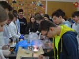 Duża grupa uczniów szkoły podstawowej stoi wzdłuż stołu laboratoryjnego. Na stole wykonują doświadczenia chemiczne.