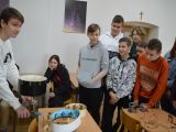 Sześcioro uczniów szkoły postawowej stoi w grupie i przygląda się jak uczeń liceum wykonuje doświadczenie z fizyki.