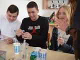 Troje uczniów szkoły podstawowej siedzi przy stoliku i przelewa płyn do naczynia.