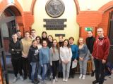 Trzymastu licealistów stoi w grupie pod ścianą. Między nimi stoją trzy kobiety.  Na ścianie portret mężczyzny i napisy.