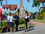 Mężczyzna w garniturze i kobieta wręczają książkę uczennicy liceum w białej bluzce.