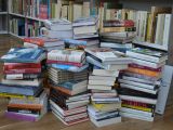 Stos książek na brązowej podłodze. W tle regał z książkami.