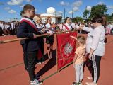 Uczeń liceum trzyma opuszczony sztandar. Po prawej stronie sztandaru stoi mały chłopiec i dziewczyna w białej bluzce, po...