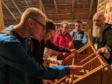 Czworo gimnazjalistów buduje makietę chaty z drewna. Obok brodaty mężczyzna w starodawnym stroju