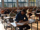 Uczniowie liceum siedzą przy stolikach uczniowskich w sali gimnastycznej. Wszyscy rozwiązują zadania na kartkach, które...