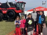 Mężczyzna z brodą i grupa uczniów szkoły podstawowej. W tle duży czerwony kombajn
